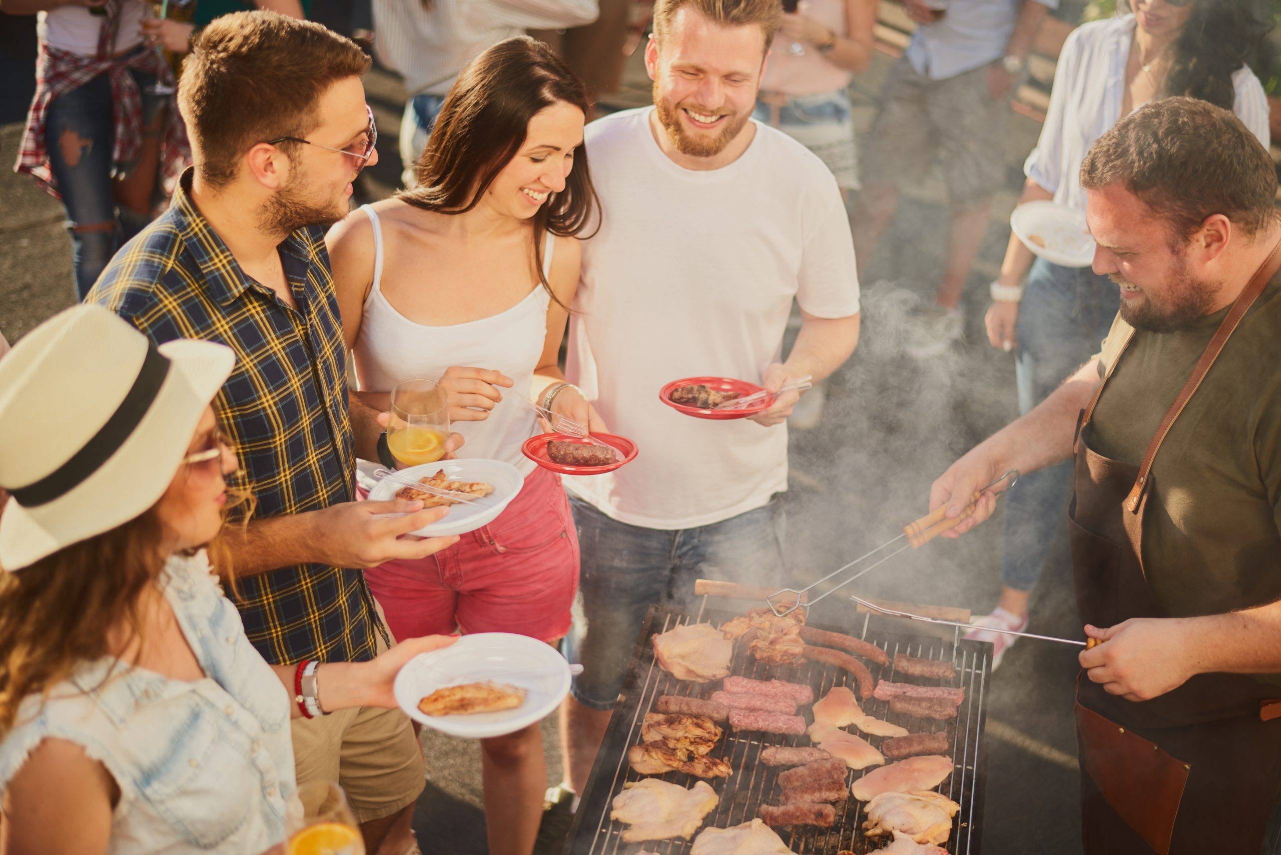 Group of people standing around grill, chatting, drinking and ea